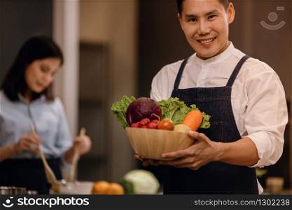 a Smiling Man with the Bowl of Fresh Vegetables, Preparing for Vegan Food in a Modern Kitchen. Blurred Woman Stirring a Healthy Salad as background. Healthy Lifestyle Concept. Cooking At Home