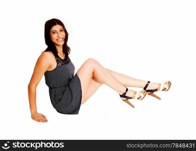 A smiling young woman in a dress sitting in profile on the floor with herlong legs, isolated on white background.