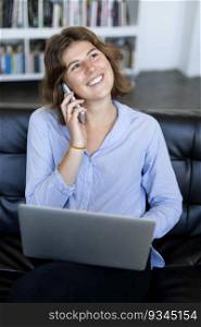 A Smiling young woman sitting on a sofa with a laptop while talking on the phone and looking at the camera.