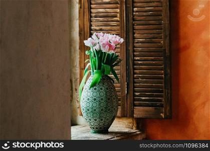 A soft focused bouquet of pink peony flowers in an old big vase on a window sill, wooden jalousie in the background, rustic style