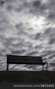 A solitary, lonely park bench in front of a sky filled with dark, moody and foreboding clouds.