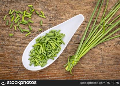 a spoon of chopped fresh chives with a bunch against rustic wood, top view