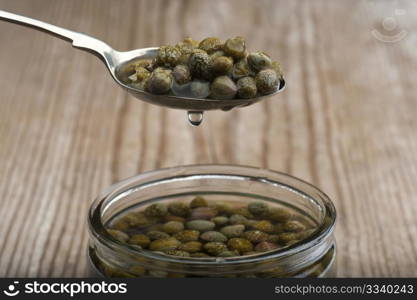A Spoonful Of Dripping Capers, Held Over Above A Jar Of Capers In Brine, On A Wooden Kitchen Table