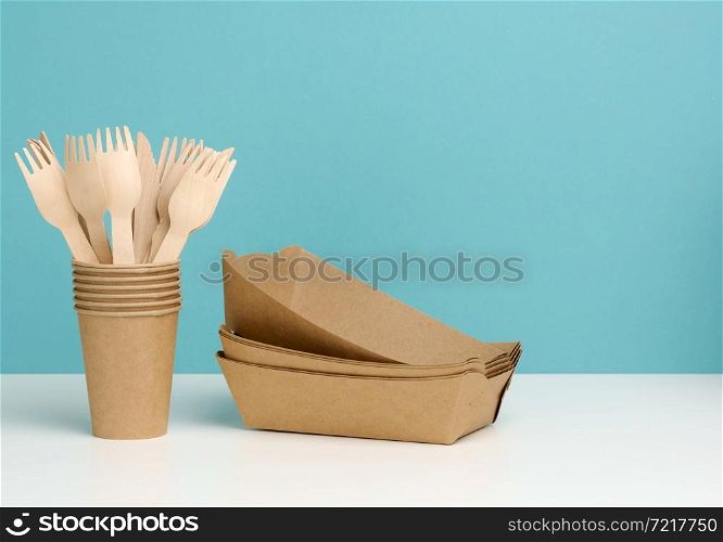 a stack of brown disposable paper cups on a white table, wooden forks and knives, blue background. Utensils for takeaway drinks, zero waste