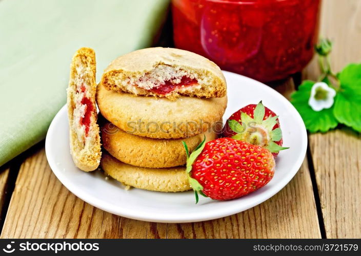 A stack of cookies filled with jam and strawberries on a plate, a jar of strawberry jam on the background of wooden boards