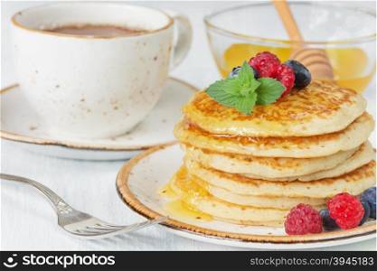 A stack of pancakes drizzled with honey and decorated with ripe raspberries and blueberries and a cup of tea