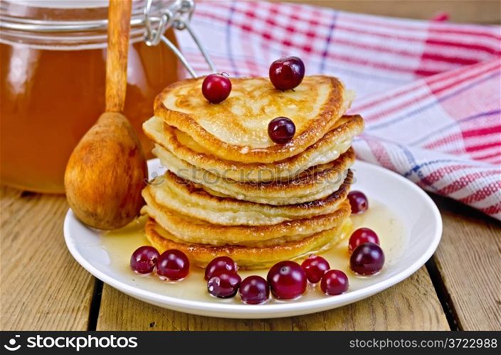 A stack of pancakes with cranberries and honey on a white plate with honey jar, a wooden spoon, napkin against a wooden board