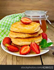 A stack of pancakes with strawberries and honey on a white plate, a jar of honey, a napkin on a wooden boards background