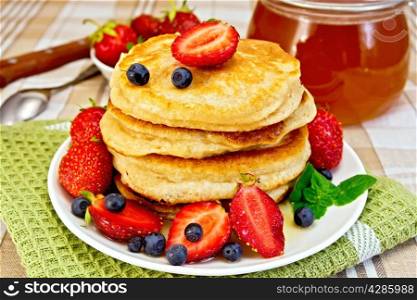A stack of pancakes with strawberries, blueberries and honey, a jar of honey, doily on linen tablecloth background