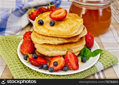 A stack of pancakes with strawberries, blueberries and honey on a white plate, jars of honey, napkins on a background of linen tablecloths