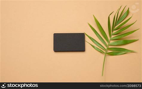 a stack of rectangular black paper business cards and a leaf of a palm tree on a brown background. View from above, modern template for branding identity