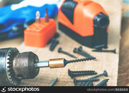 A steel screw is placed on an electric drill on a wooden board background. The concept of tools and repair work. Steel screws. Metal screw. A steel screw is placed on an electric drill on a wooden board background. The concept of tools and repair work. Steel screws.