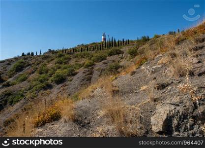 A steep rocky slope with slender cypresses and a lighthouse on a sunny summer day, Crimea.