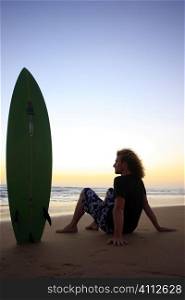 A stock photograph of a young man sitting on the beach with his surfboard watching the sunset.