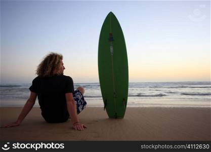 A stock photograph of a young man sitting on the beach with his surfboard watching the sunset.