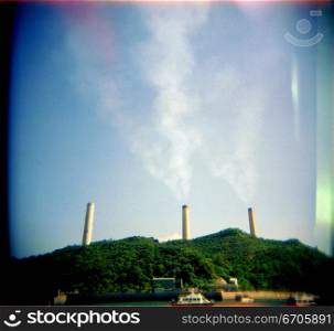 A stock photograph of pollution coming from tall chimneys behind a green hill, using a Holga camera which acheives an artistic and photographic effect.