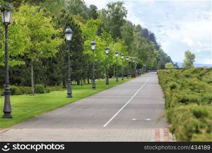 A straight asphalt road and a bike path go along a beautiful, clipped green lawn through a summer park, on the edge of the road are many beautiful old lamps.. Straight asphalt road and bike path through the park with many beautiful vertical vintage lights