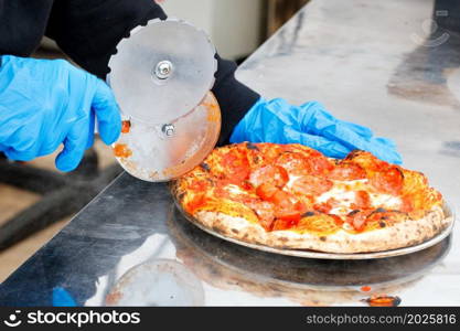 A street chef in blue latex gloves cuts an oven-baked pizza with a round knife. Copy space.. The hands of the cook cut the pizza cooked in the oven with a round knife.