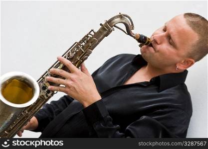A street musician leaning against a white wall and playing his saxophone.