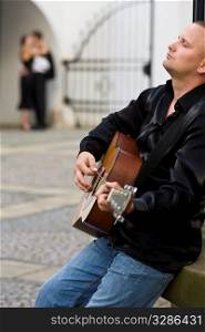A street musician playing his guitar serenades a romantic couple kissing out of focus in the background