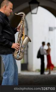 A street musician plays his saxophone while a romantic couple can be seen out of focus in the background
