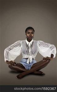 A studio portrait of a serene young black female sitting on the floor with short black hair and pink nails wearing a white sheer blouse with denim shorts and silver jewelry.