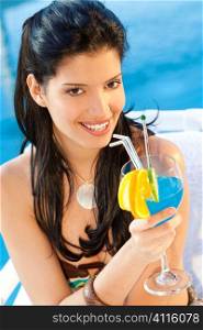 A stunningly beautiful young hispanic woman looking drinking a cocktail next to a swimming pool
