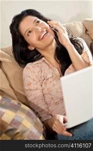 A stunningly beautiful young Latin Hispanic woman sitting on a settee having fun on her laptop and laughing while talking on her cell phone