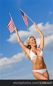 A stunningly beautiful young woman in a white bikini holds two stars and stripes flags above her head