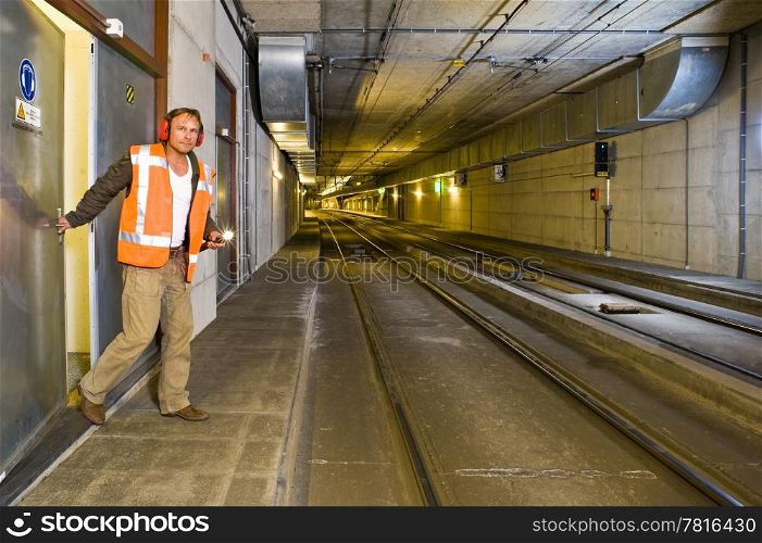 A sturdy looking engineer, closing a door to a maintance room behind him inside a public transportation tunnel, looking relaxed and with a flashlight in his hand