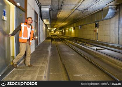 A sturdy looking engineer, closing a door to a maintance room behind him inside a public transportation tunnel, looking relaxed and with a flashlight in his hand