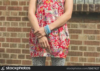 A stylish young woman wearing a colorful dress and bracelets is standing by a red brick wall