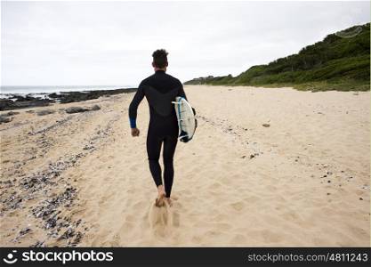 A surfer dude walks along the beach with a surfboard tuck underneath his arm while wearing a wetsuit as seen from behind.