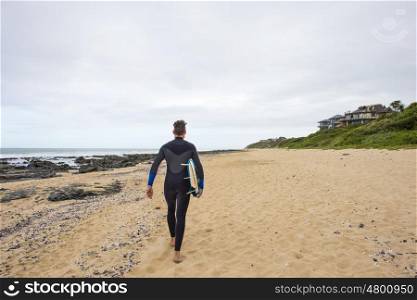A surfer in a wetsuit walks along the beach with his surfboard under his arm.