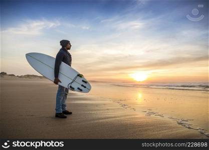 A surfer with his surfboard at the sunset looking to the waves