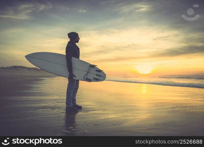 A surfer with his surfboard at the sunset looking to the waves