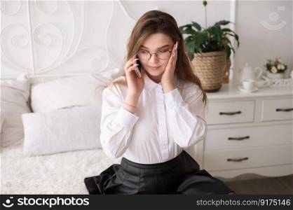 A surprised emotional European girl in a white shirt and glasses is talking on the phone. Sitting on a white bed. Hand on the face. Working day at home. Emotional women with glasses talking on the phone and getting upset