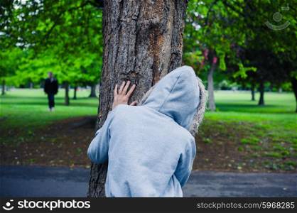 A suspicious character wearing a hoodie is hiding behind a tree in the park
