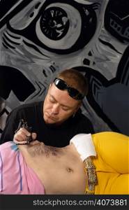 A tattoo artist applying his craft onto the abdomen of a female. (Property release supplied includes tattooists&acute; wall mural behind him.)