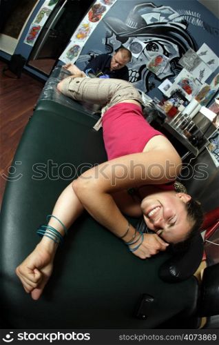 A tattoo artist applying his craft onto the lower leg of a pain-inflicted female. (Property release supplied includes tattooists&acute; wall mural and drawings posted on the walls behind him.)