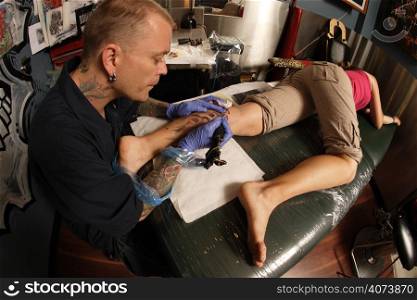 A tattoo artist applying his craft onto the lower leg of a pain-inflicted female. (Property release supplied includes tattooists&acute; wall mural and drawings posted on the walls behind him.)