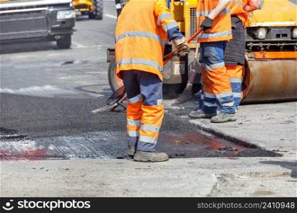 A team of road workers in orange overalls patch up part of the road with fresh asphalt, using shovels, levels, a broom and road equipment to repair a small section of the road. Copy space.. A group of road workers in orange overalls are repairing a section of the carriageway by rolling up fresh asphalt.