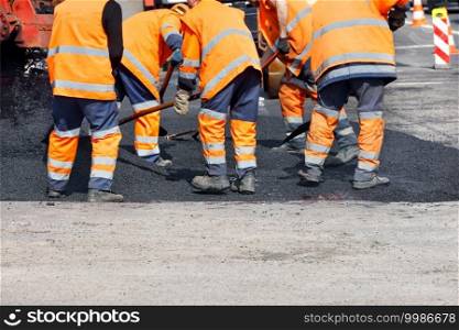 A team of road workers in orange reflective uniforms cover a section of road with fresh asphalt to repair the section of the road. Clear day, copy space.. A team of road workers in orange reflective uniforms use shovels to level fresh asphalt on a clear day.