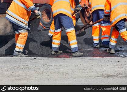A team of road workers in orange uniforms resurfaces part of the road with fresh asphalt, spreading it with shovels, then compacting it with a road roller. Copy space.. A road worker is spreading fresh asphalt with shovels over the repair area to repair a section of road.