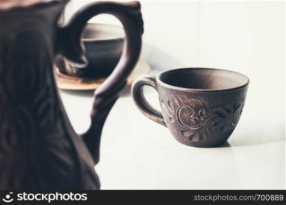 A teapot in the foreground and a cup of clay stand near the window. Close-up.. A teapot in the foreground and a cup of clay stand near the window.