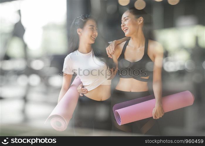 A teenage girl friend is walking holding a purple yoga mats after giving up yoga on the gym blurry foreground, A photo through a glass window of a young Asian woman talking and laughing at a fitness facility.