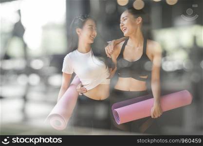 A teenage girl friend is walking holding a purple yoga mats after giving up yoga on the gym blurry foreground, A photo through a glass window of a young Asian woman talking and laughing at a fitness facility.