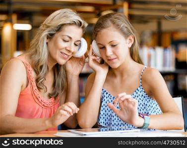 A teenage girl with headphones sitting in a library with her mum. That is fun wth mum