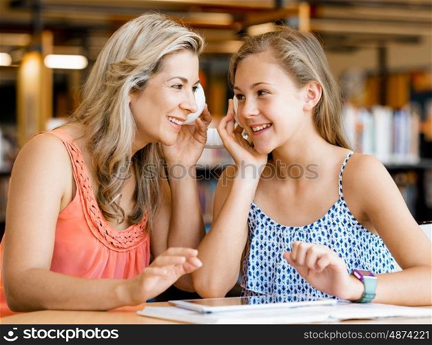 A teenage girl with headphones sitting in a library with her mum. That is fun wth mum