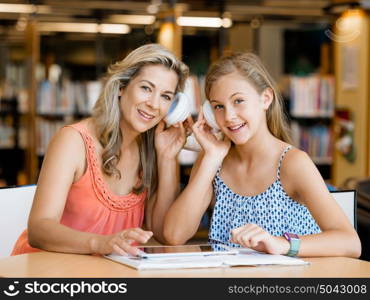 A teenage girl with headphones sitting in a library with her mum. That is fun wth mum